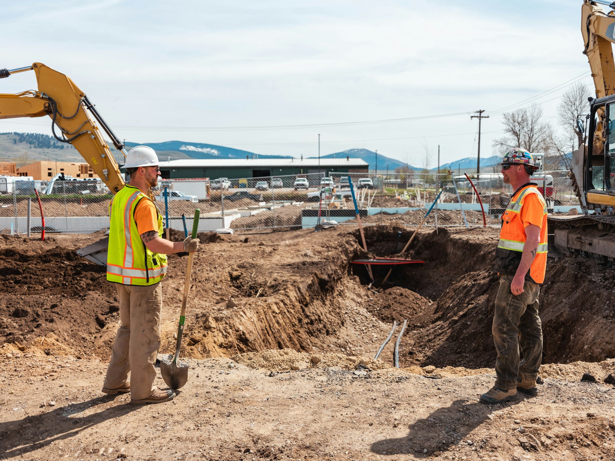 Two crew members in hard hats and high visibility vests at a utility trench with an excavator behind them