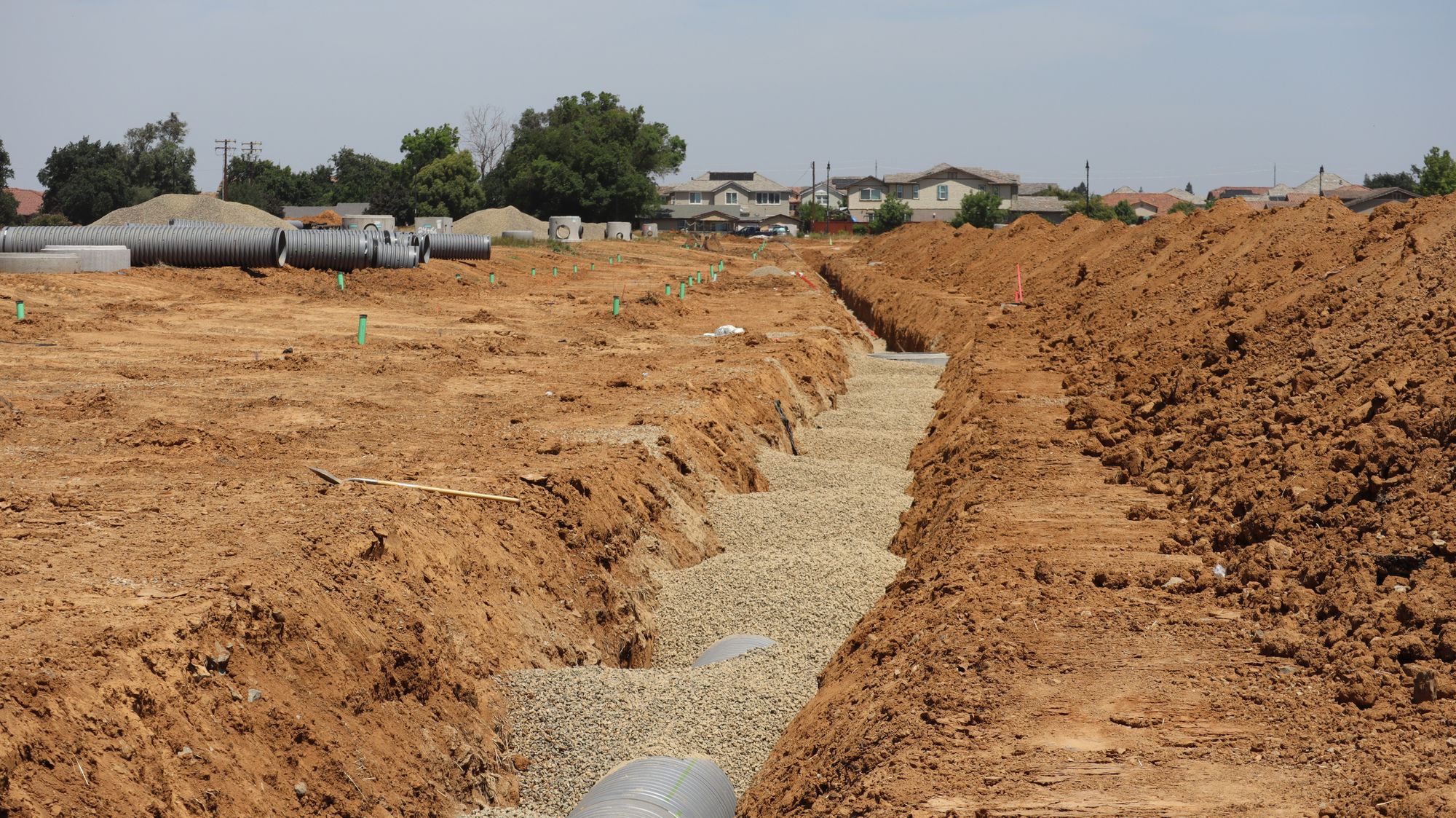 Open utility trench with pipe laid in a gravel bed next to a new residential neighborhood